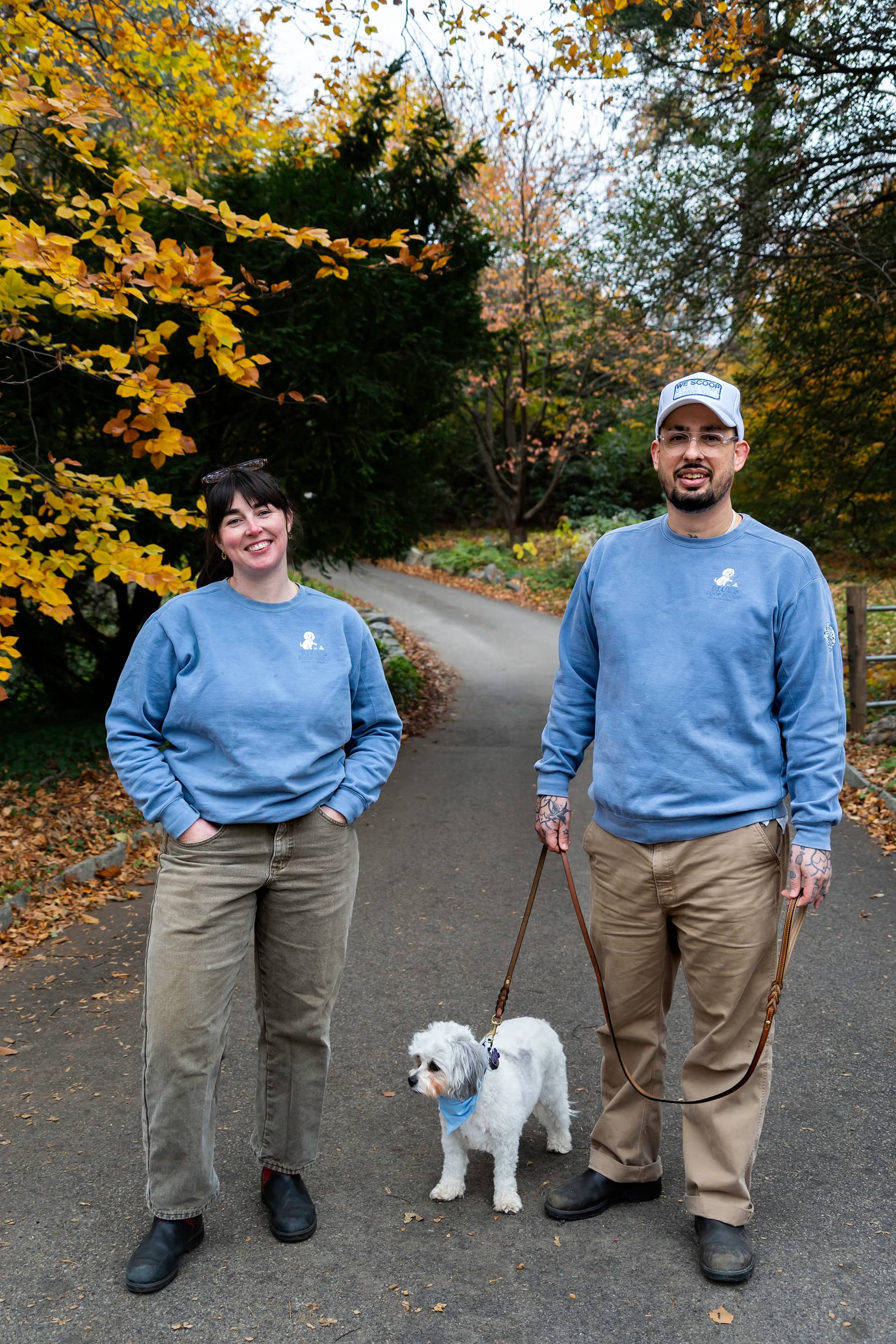 Both owners in branded sweatshirts on a leafy path, their small white dog on a lead